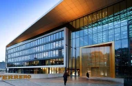 A imagem mostra um prédio com fachada de vidro e detalhes dourados, onde está escrito “FERRERO” em letras grandes na entrada. Pessoas caminham em frente ao edifício, que reflete o céu azul e árvores ao redor