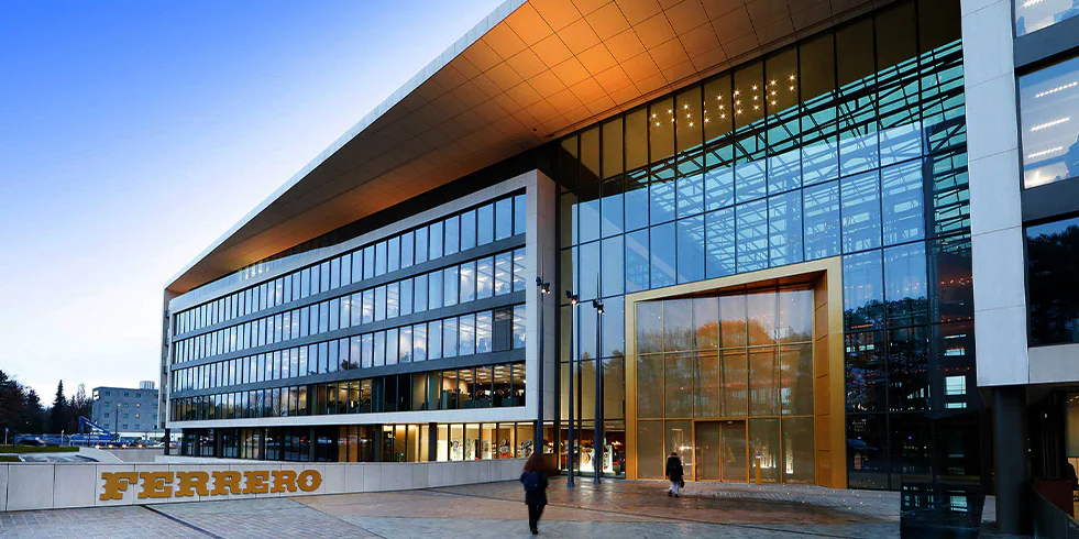A imagem mostra um prédio com fachada de vidro e detalhes dourados, onde está escrito “FERRERO” em letras grandes na entrada. Pessoas caminham em frente ao edifício, que reflete o céu azul e árvores ao redor