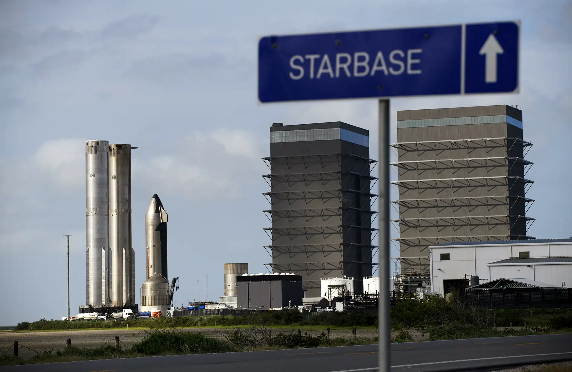 A imagem exibe uma área industrial com uma placa azul escrita "STARBASE" e uma seta para cima. Ao fundo, há três estruturas metálicas cilíndricas, semelhantes a componentes de foguetes, e dois edifícios altos com laterais cinza. Equipamentos e veículos industriais estão em destaque em um ambiente amplo de lançamento espacial.