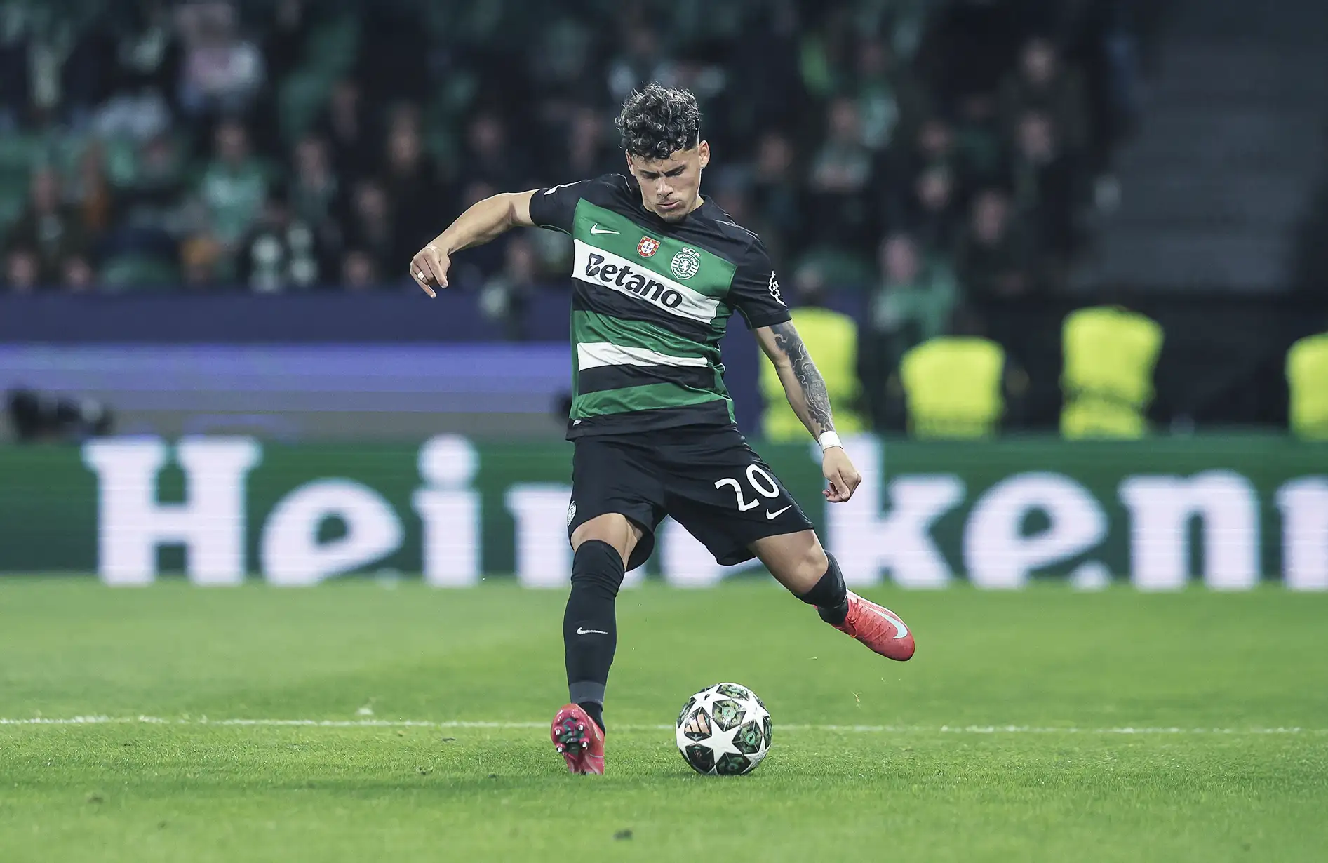 A imagem mostra um jogador de futebol em um campo, vestindo uniforme listrado verde e preto, shorts e meias pretas. Ele está prestes a chutar uma bola, com o pé direito levantado. Ao fundo, há espectadores e uma placa publicitária da Heineken em um estádio.
