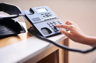 A imagem mostra uma mesa de escritório com um telefone fixo preto em destaque, que possui um visor digital e botões numéricos. Uma mão está pressionando um dos botões, enquanto documentos estão organizados em um suporte ao lado, sugerindo um ambiente de trabalho bem iluminado.