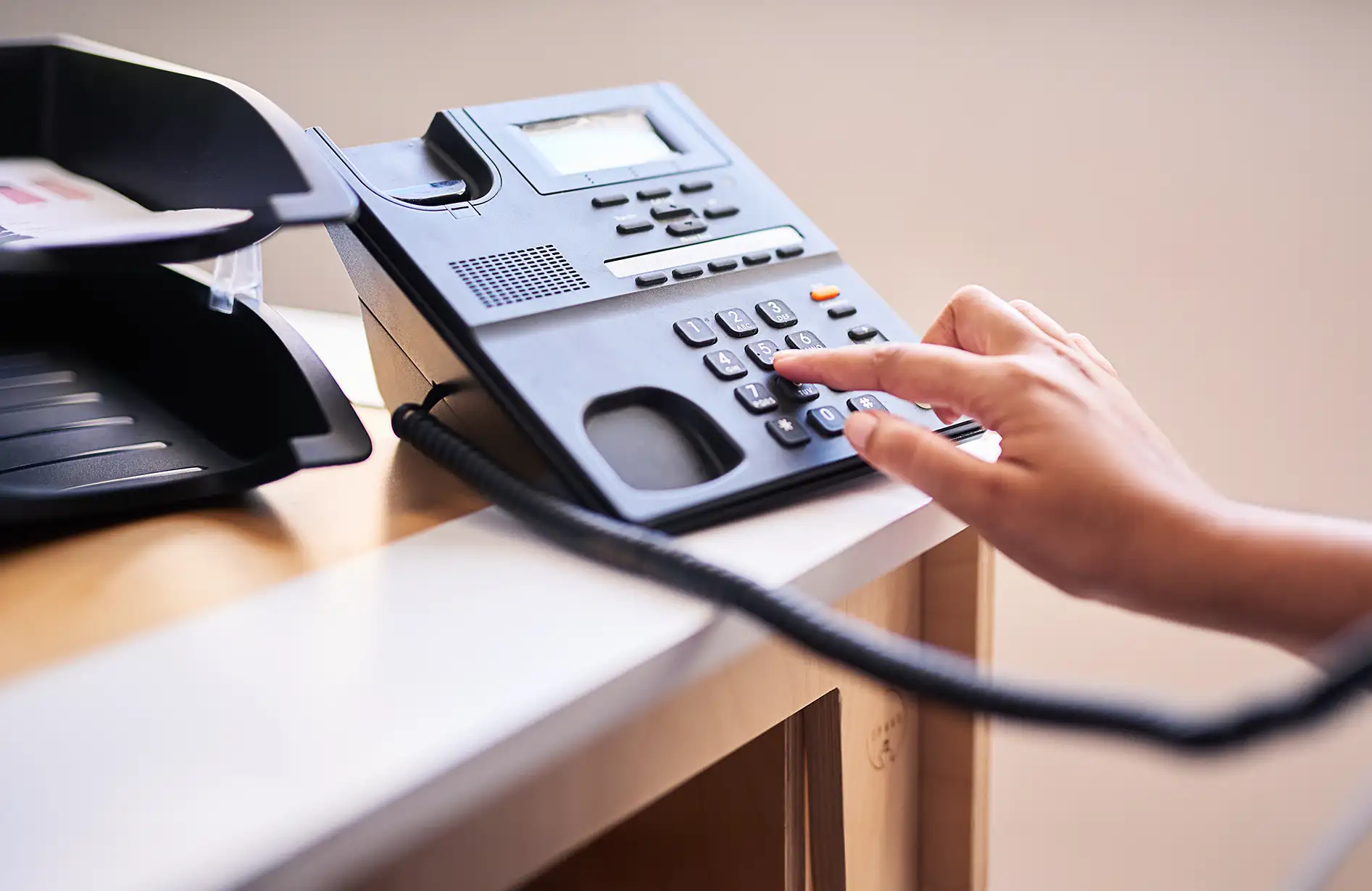 A imagem mostra uma mesa de escritório com um telefone fixo preto em destaque, que possui um visor digital e botões numéricos. Uma mão está pressionando um dos botões, enquanto documentos estão organizados em um suporte ao lado, sugerindo um ambiente de trabalho bem iluminado.