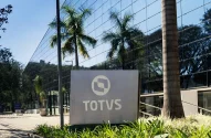 A imagem mostra a fachada de um prédio moderno com vidro refletindo o céu azul, cercado por árvores. Em frente, um letreiro exibe o logotipo da Totvs. Há uma área com vegetação ornamental, palmeiras e um caminho pavimentado até a entrada do edifício.