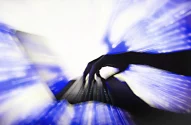 A imagem mostra uma mão em silhueta sobre um teclado de computador preto com iluminação sob as teclas. O fundo, desfocado em azul e branco, cria uma sensação de movimento e dinamismo, sugerindo rapidez na digitação ou interatividade digital, com padrões de luz evocando energia ou fluxo de dados.