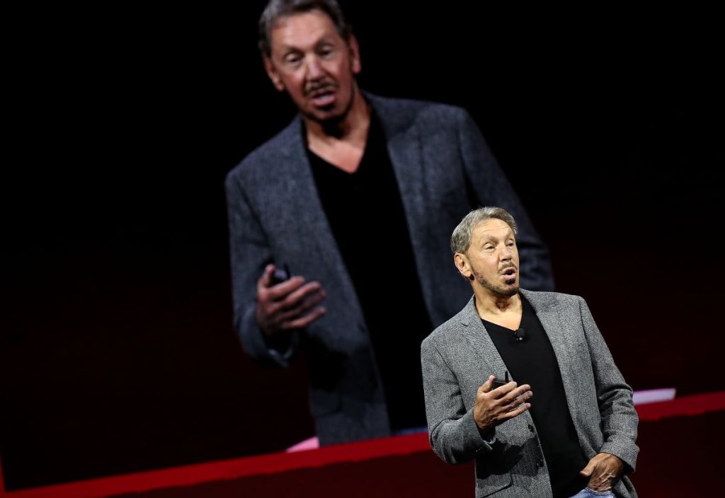 Fundador da Oracle está de paletó cinza e camiseta preta dando uma palestra