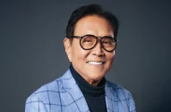 A imagem mostra Robert Kiyosaki, escritor do best-seller “Pai Rico, Pai Pobre”: um homem sorridente de óculos, blazer xadrez azul claro e camisa preta de gola alta, sobre fundo cinza neutro.
