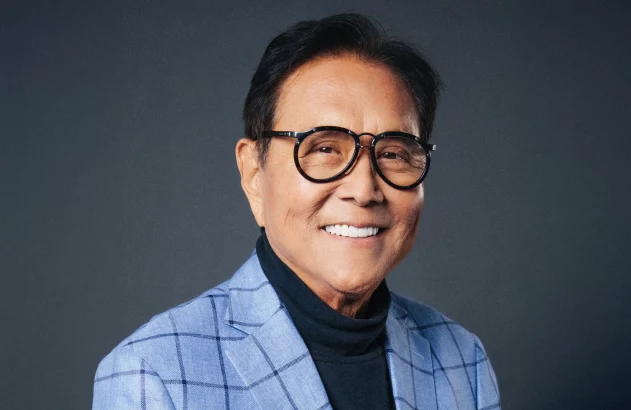 A imagem mostra Robert Kiyosaki, escritor do best-seller “Pai Rico, Pai Pobre”: um homem sorridente de óculos, blazer xadrez azul claro e camisa preta de gola alta, sobre fundo cinza neutro.