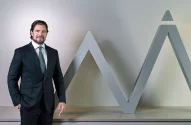 Homem de terno escuro e gravata posa sorrindo ao lado de uma estrutura metálica cinza em forma de “M” com topo triangular.