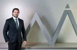 Homem de terno escuro e gravata posa sorrindo ao lado de uma estrutura metálica cinza em forma de “M” com topo triangular.