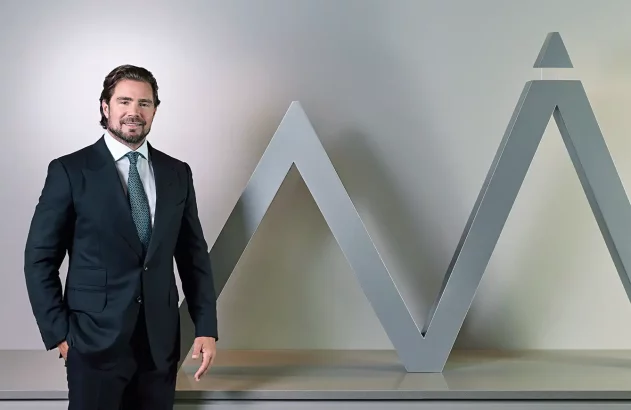 Homem de terno escuro e gravata posa sorrindo ao lado de uma estrutura metálica cinza em forma de “M” com topo triangular.