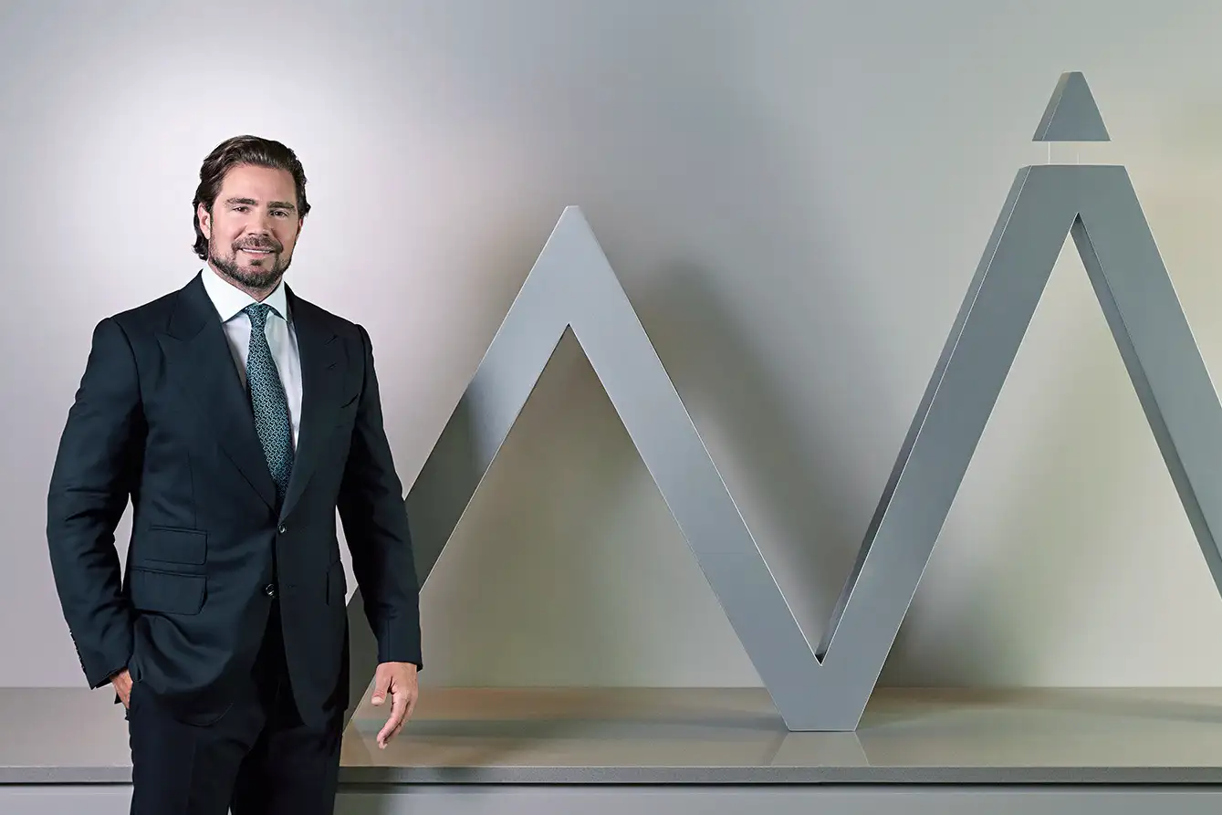 Homem de terno escuro e gravata posa sorrindo ao lado de uma estrutura metálica cinza em forma de “M” com topo triangular.