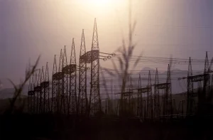 Torres de transmissão de energia se estendem até o horizonte ao entardecer, destacando-se em silhuetas contra um céu lilás e laranja, com montanhas ao fundo.