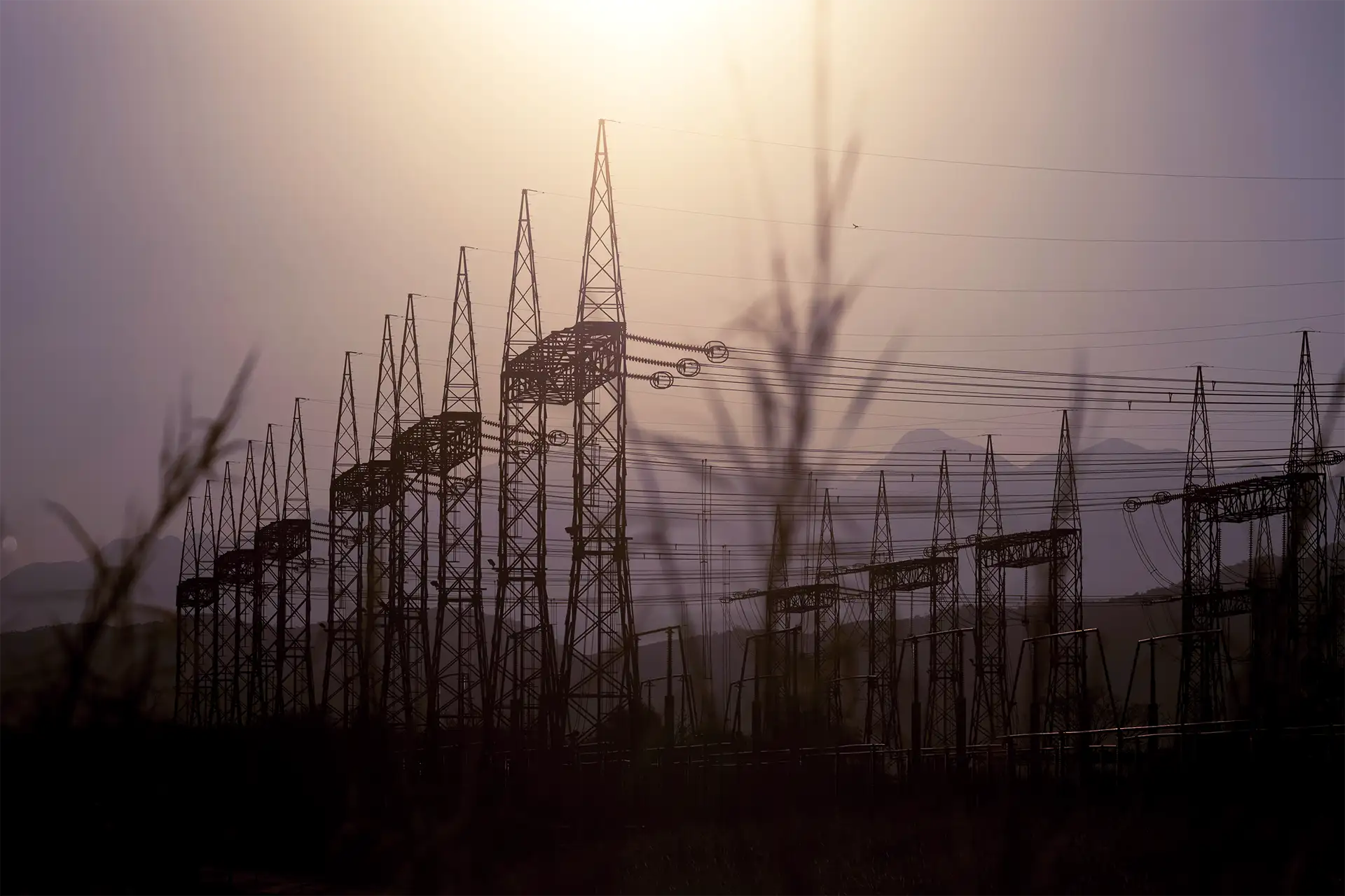 Torres de transmissão de energia se estendem até o horizonte ao entardecer, destacando-se em silhuetas contra um céu lilás e laranja, com montanhas ao fundo.