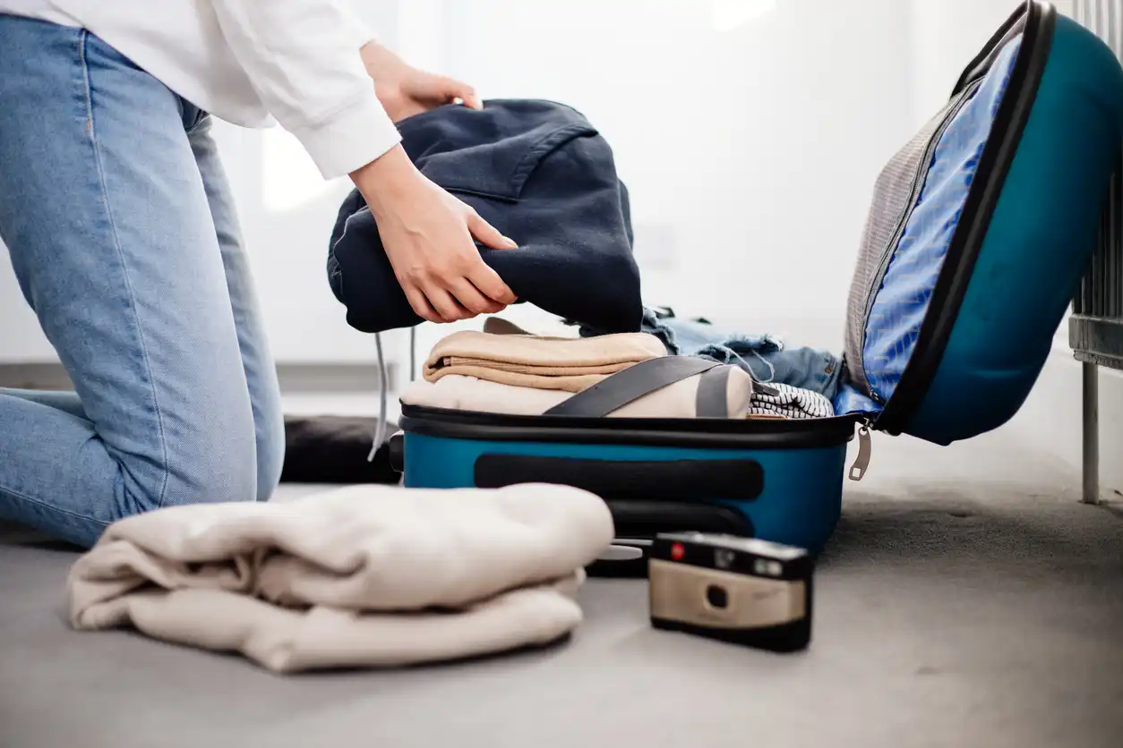 Pessoa fazendo as malas para uma viagem, organizando roupas e objetos em uma mala de viagem azul.