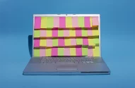 A imagem mostra um laptop prata aberto em um fundo azul, com a tela coberta por notas adesivas rosa, verde-limão e amarelo, formando um padrão colorido.