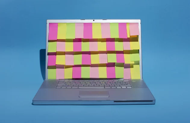 A imagem mostra um laptop prata aberto em um fundo azul, com a tela coberta por notas adesivas rosa, verde-limão e amarelo, formando um padrão colorido.