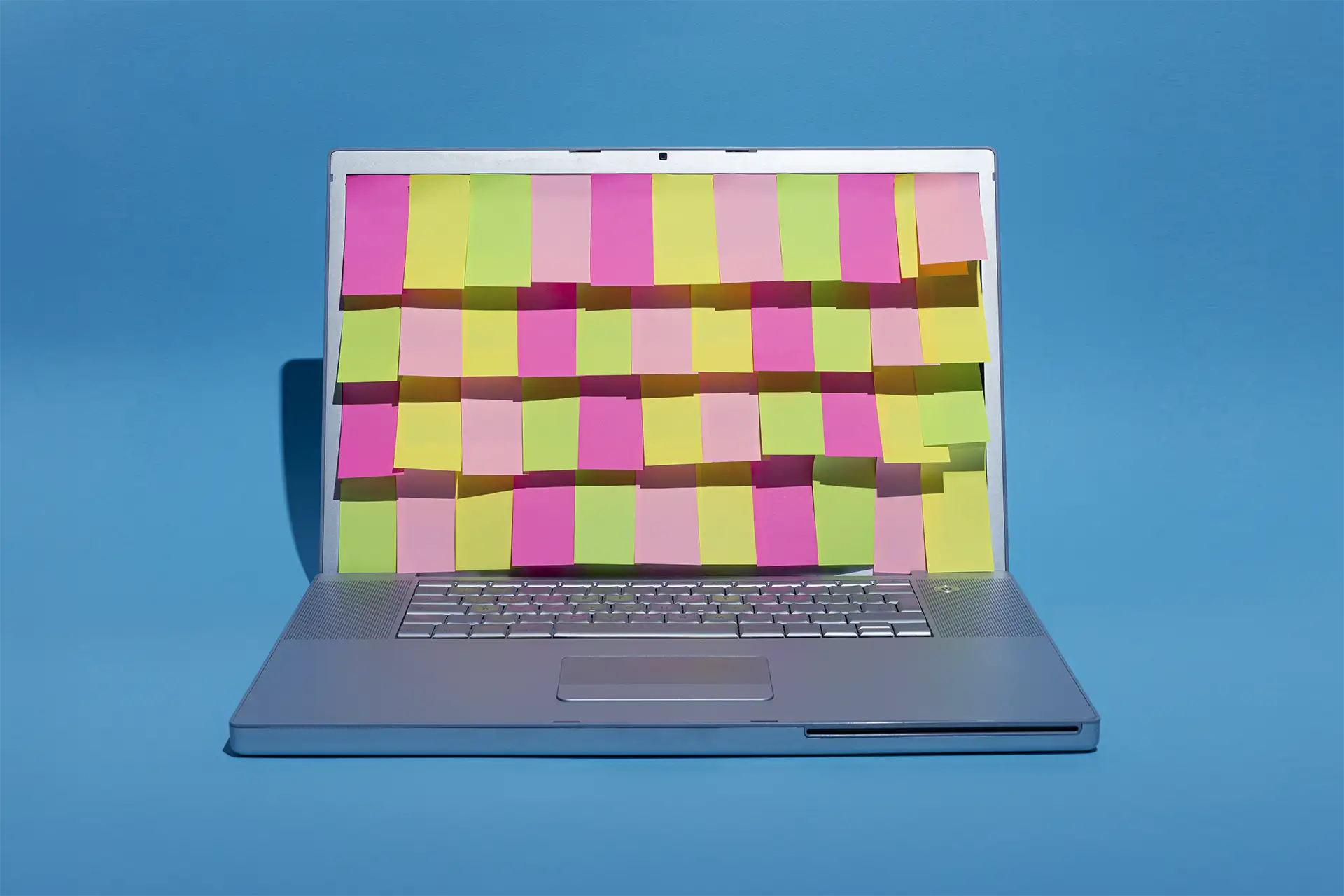 A imagem mostra um laptop prata aberto em um fundo azul, com a tela coberta por notas adesivas rosa, verde-limão e amarelo, formando um padrão colorido.