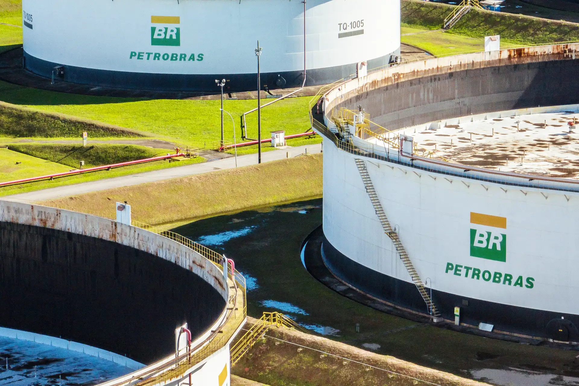 A imagem mostra dois tanques brancos de armazenamento da Petrobras em uma área externa gramada, cercados, com tubulações vermelhas e postes de iluminação.