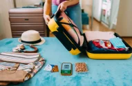 Pessoa preparando uma bolsa de viagem com itens essenciais como remédios, máscara, cartão de embarque, e celular, em uma mesa com roupas e acessórios.