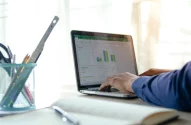 Pessoa trabalhando em computador portátil com gráficos e planilhas abertas, em ambiente bem iluminado.