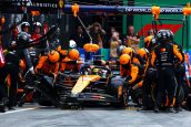 Imagem da equipe da MaLaren de Formula 1, com engenheiros de macacão preto e laranja em volta de um carro de corrida