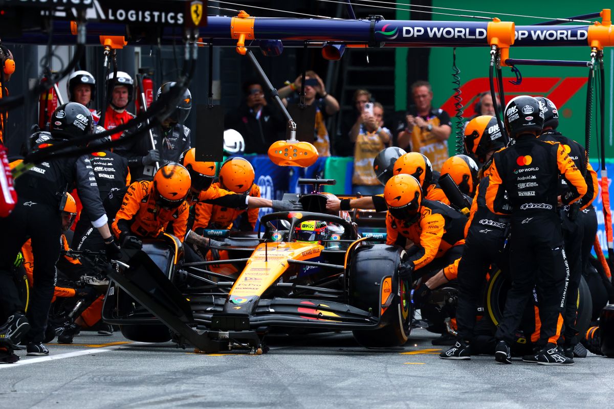 Imagem da equipe da MaLaren de Formula 1, com engenheiros de macacão preto e laranja em volta de um carro de corrida