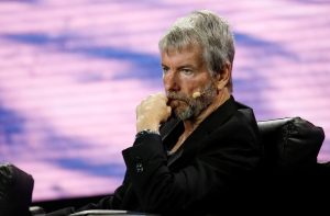 Foto de Michael Saylor, da Strategy: um senhor grisalho, magro, de olhos azuis, barba cerrada, olha para frente e segura a boca com a mão esquerda, está de terno preto e relógio prateado