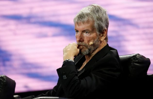 Foto de Michael Saylor, da Strategy: um senhor grisalho, magro, de olhos azuis, barba cerrada, olha para frente e segura a boca com a mão esquerda, está de terno preto e relógio prateado