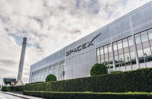 Fachada da SpaceX, com letras cinzas em um prédio baixo branco, céu está cheio de nuvens