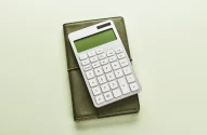 Calculadora branca sobre caderno verde, em fundo claro, com composição minimalista e iluminação suave.