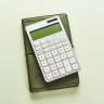 Calculadora branca sobre caderno verde, em fundo claro, com composição minimalista e iluminação suave.