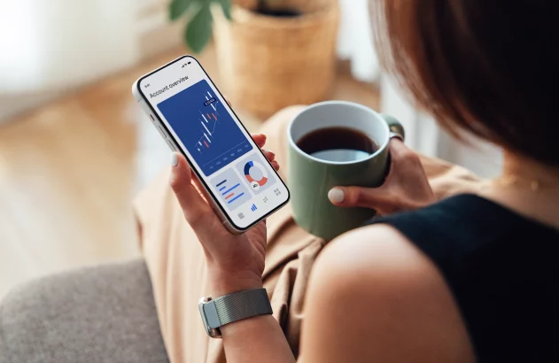 Mulher segura celular com gráficos de investimentos e xícara de café, simbolizando controle financeiro e investimentos pessoais.