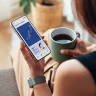 Mulher segura celular com gráficos de investimentos e xícara de café, simbolizando controle financeiro e investimentos pessoais.