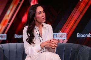 Cristina Junqueira, do Nubank, está com uma roupa branca, cabelos pretos soltos, sentada em um sofá cinza