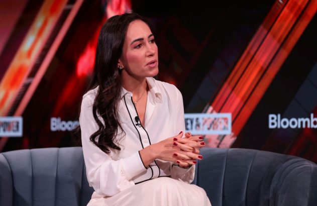 Cristina Junqueira, do Nubank, está com uma roupa branca, cabelos pretos soltos, sentada em um sofá cinza