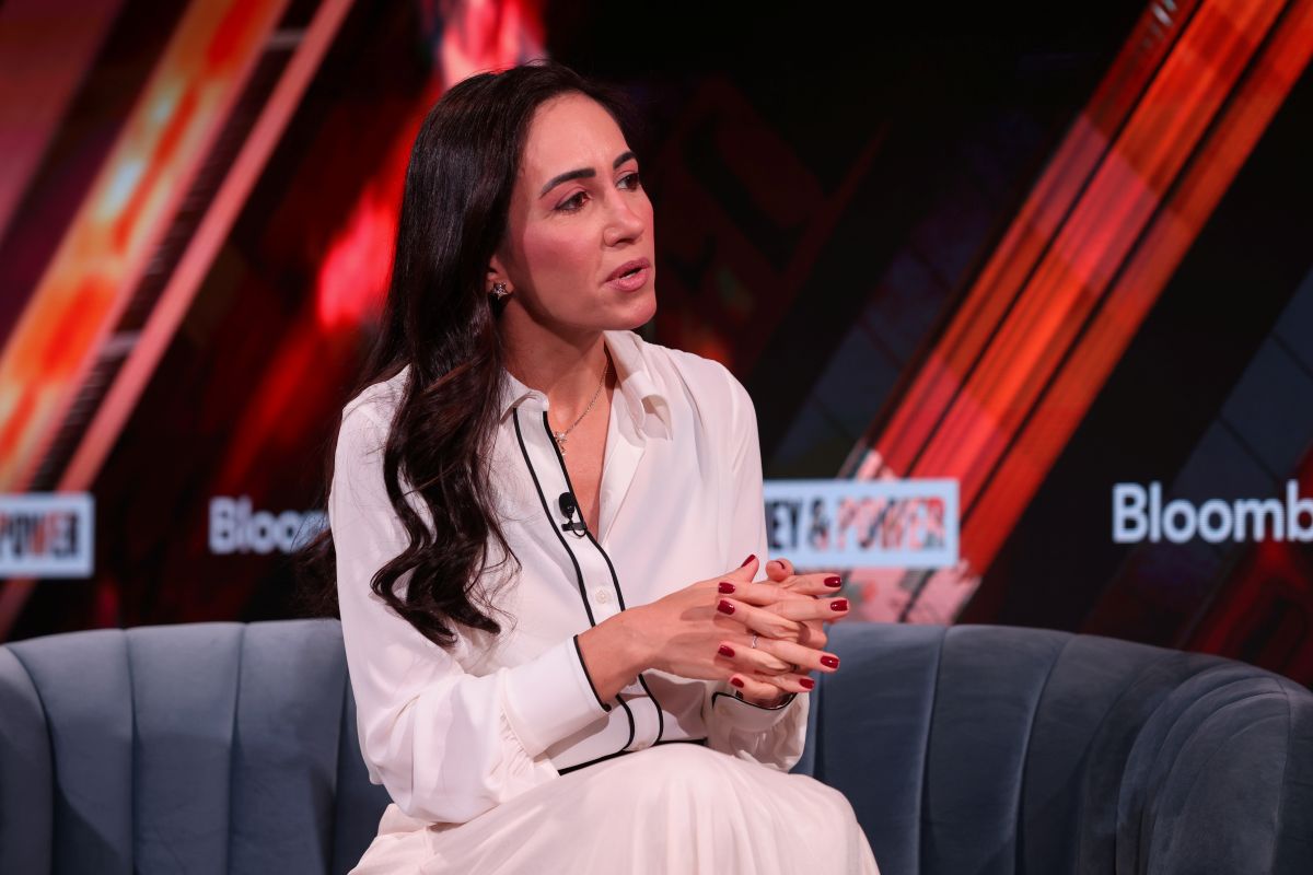 Cristina Junqueira, do Nubank, está com uma roupa branca, cabelos pretos soltos, sentada em um sofá cinza