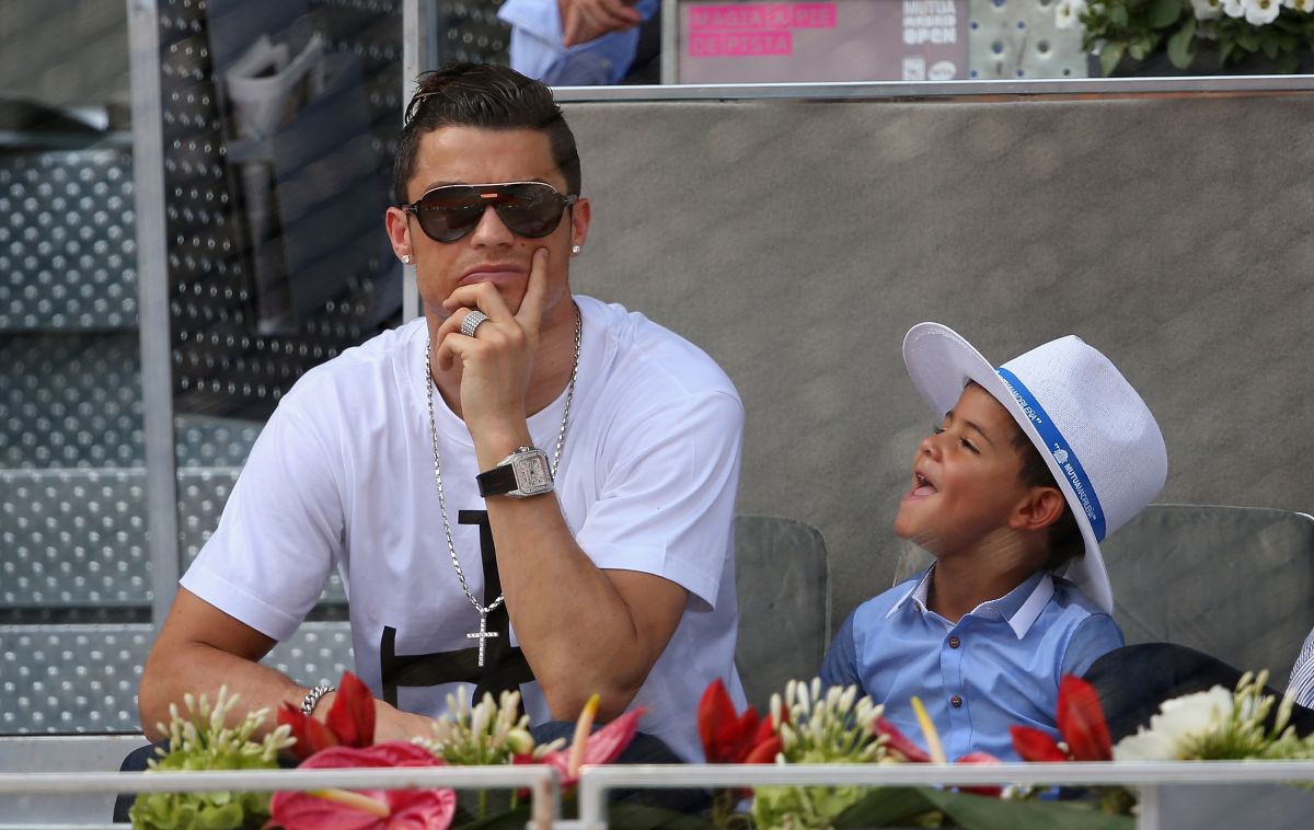 Cristiano Ronaldo está vendo um jogo, de camisa branca, óculos escuros, relógio e colar de ouro