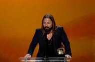 Max Martin, produtor de Taylor Swift, tem cabelos lisos e longos e loiros, olhos azuis, está com um blazer preto em frente a uma parede laranja