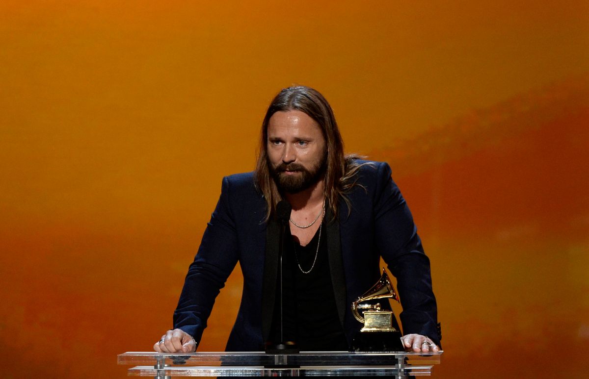 Max Martin, produtor de Taylor Swift, tem cabelos lisos e longos e loiros, olhos azuis, está com um blazer preto em frente a uma parede laranja