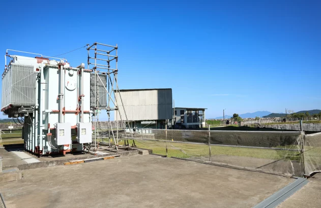 Usina nuclear em complexo industrial costeiro, com reatores, subestação e píer, cercada por floresta e montanhas.