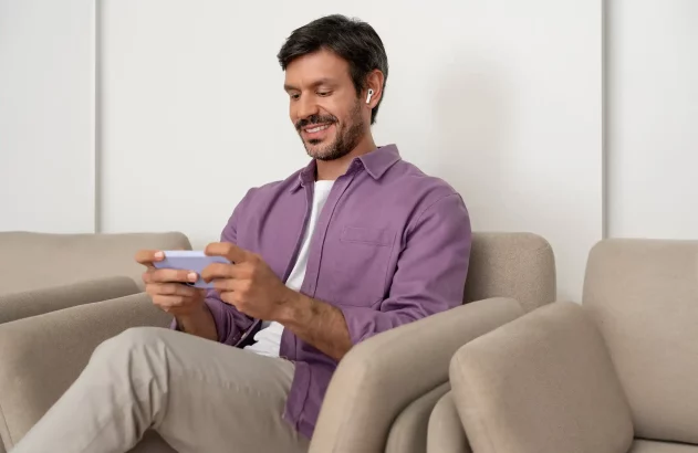 Homem sentado em sofá segurando celular com as duas mãos e usando fones de ouvido sem fio.