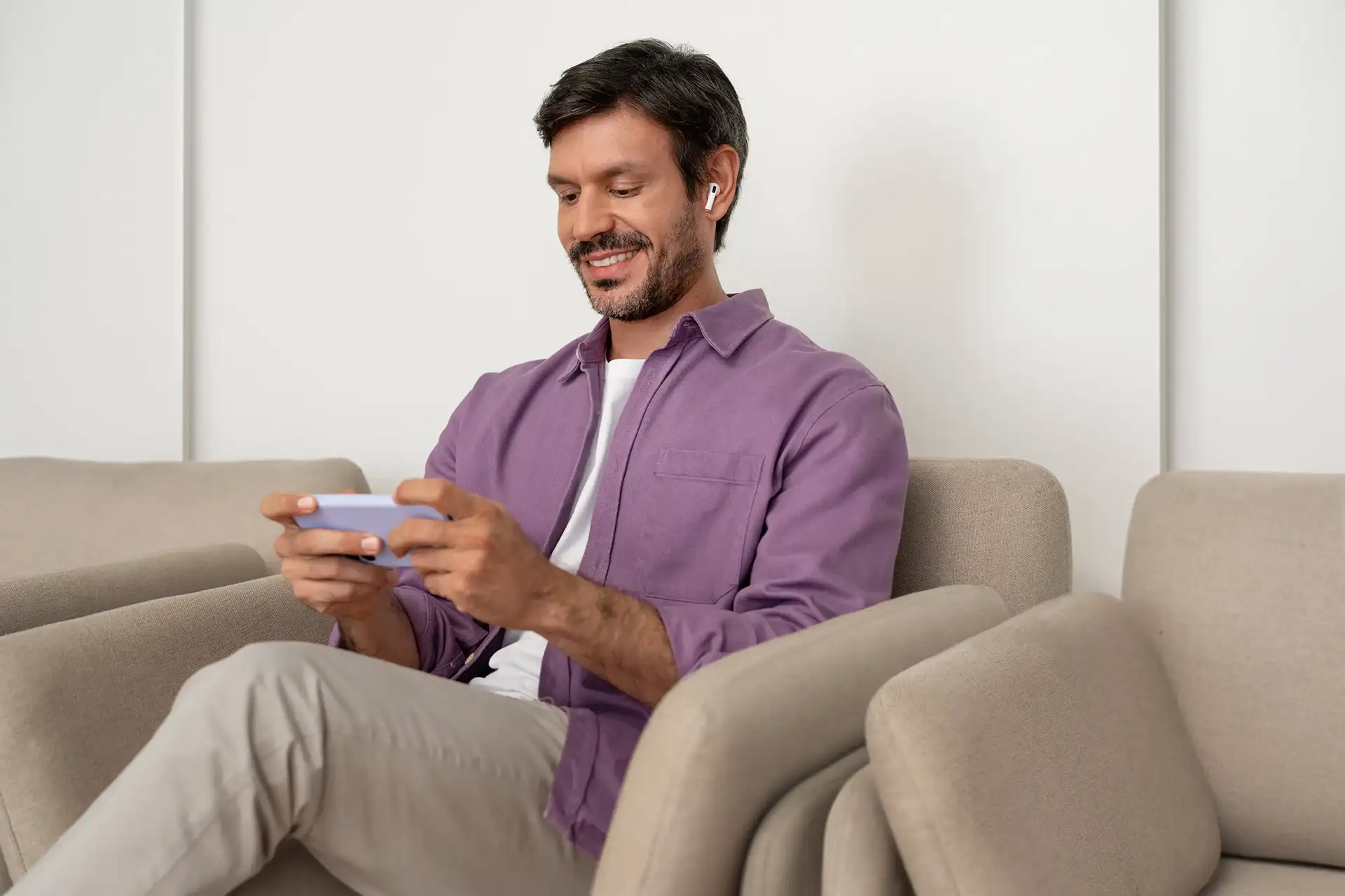 Homem sentado em sofá segurando celular com as duas mãos e usando fones de ouvido sem fio.
