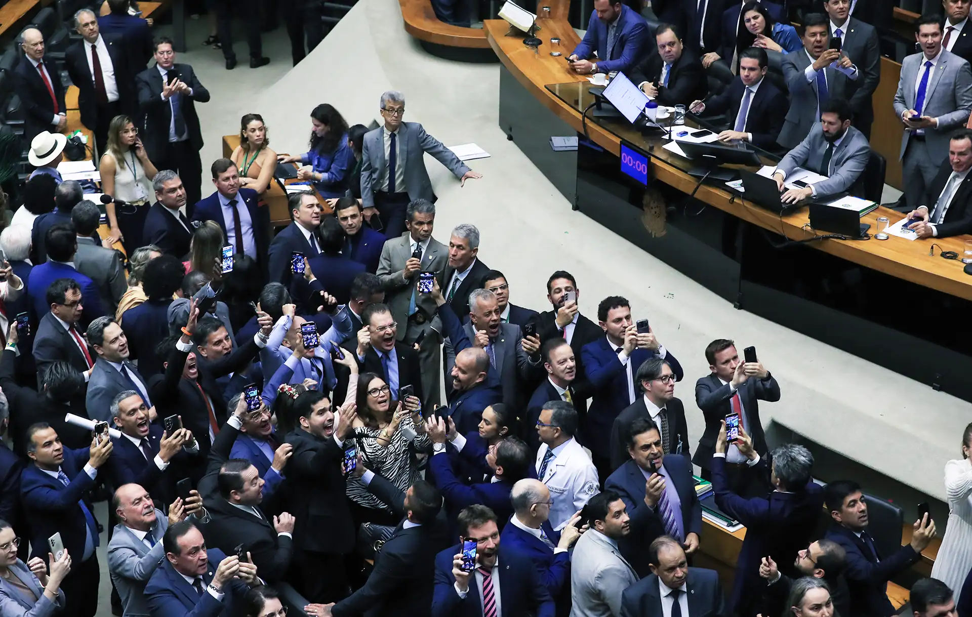 Plenário lotado com dezenas de parlamentares sorrindo e levantando celulares para registrar a comemoração após votação.