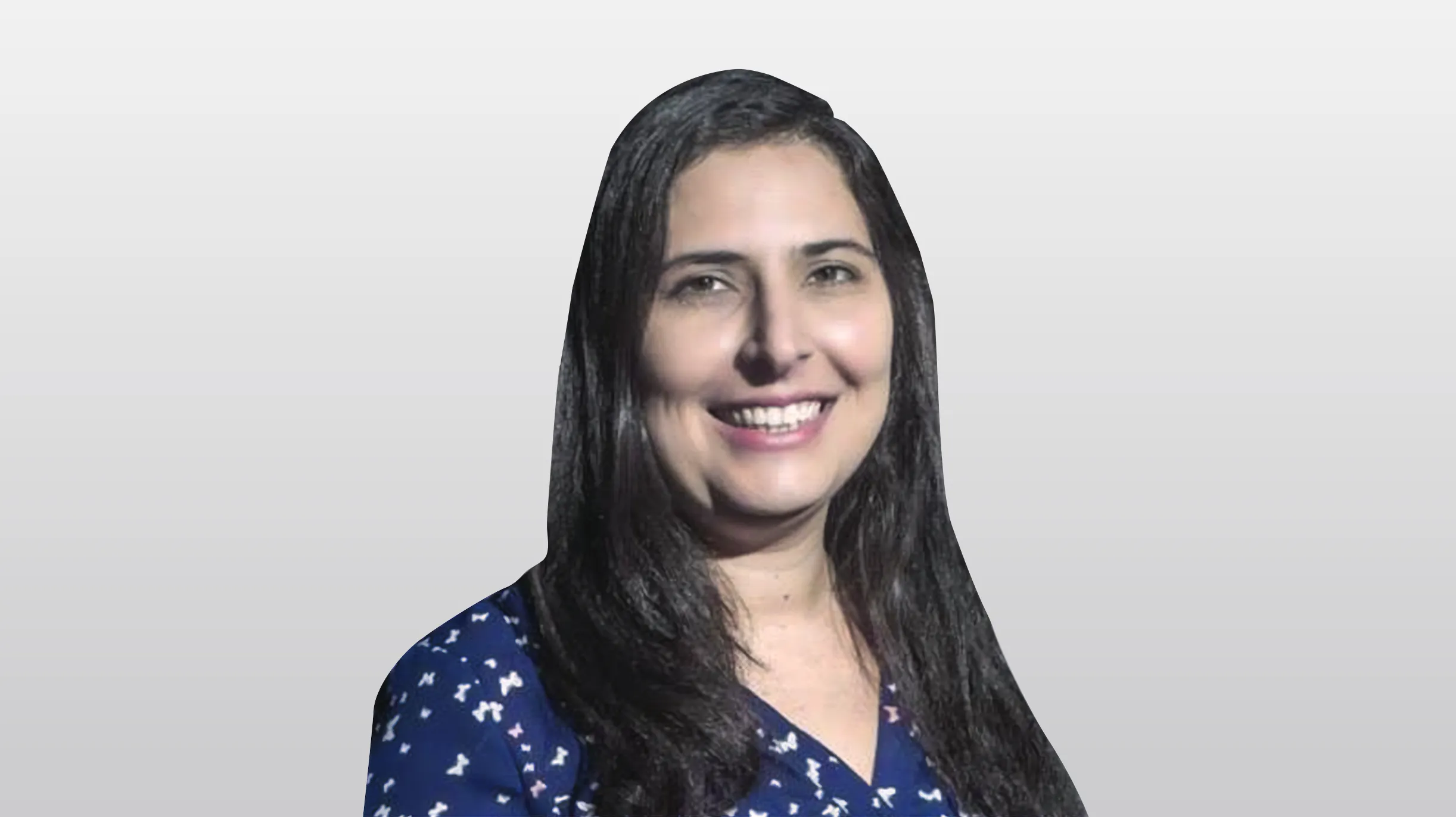 Retrato de uma mulher de pele clara e longos cabelos escuros, vestindo uma blusa azul marinho com estampa com detalhes brancos. Ela sorri, mostrando os dentes, em um fundo branco e liso.