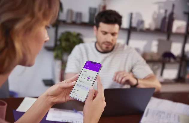 A imagem apresenta uma cena onde duas pessoas estão em um ambiente interno, como um escritório ou sala de estar. Em primeiro plano, uma mulher segura um smartphone mostrando um aplicativo de banco em um fundo roxo. Ao fundo, um homem trabalha concentrado em um laptop, cercado por papéis, sugerindo uma atividade administrativa. O ambiente é iluminado suavemente, com uma estante de livros e uma planta que adicionam um ar acolhedor e organizado.