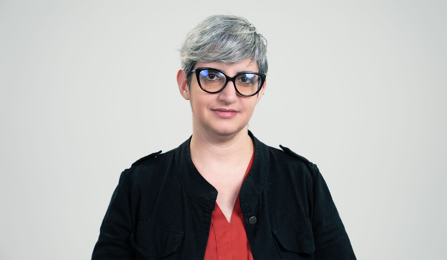 Retrato de mulher branca de cabelos grisalhos curtos vestindo óculos de armação preta, blusa vermelha e jaqueta preta.