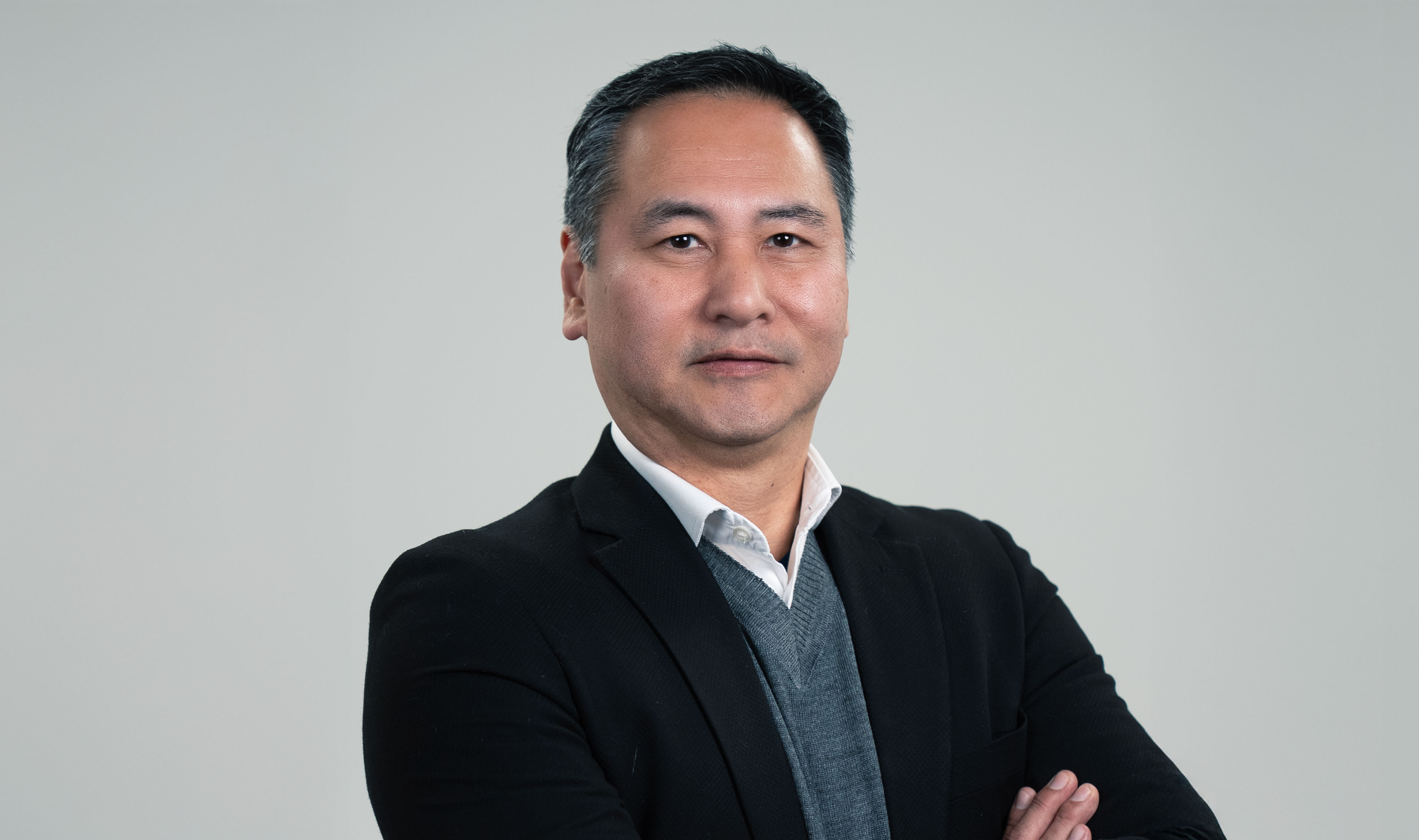 Retrato de homem asiático vestindo camisa branca, suéter cinza e terno preto.
