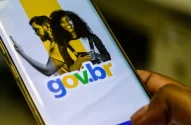 Smartphone com app gov.br aberto. Mão posiciona dedo para tocar botão "Entrar com gov.br". imposto de renda