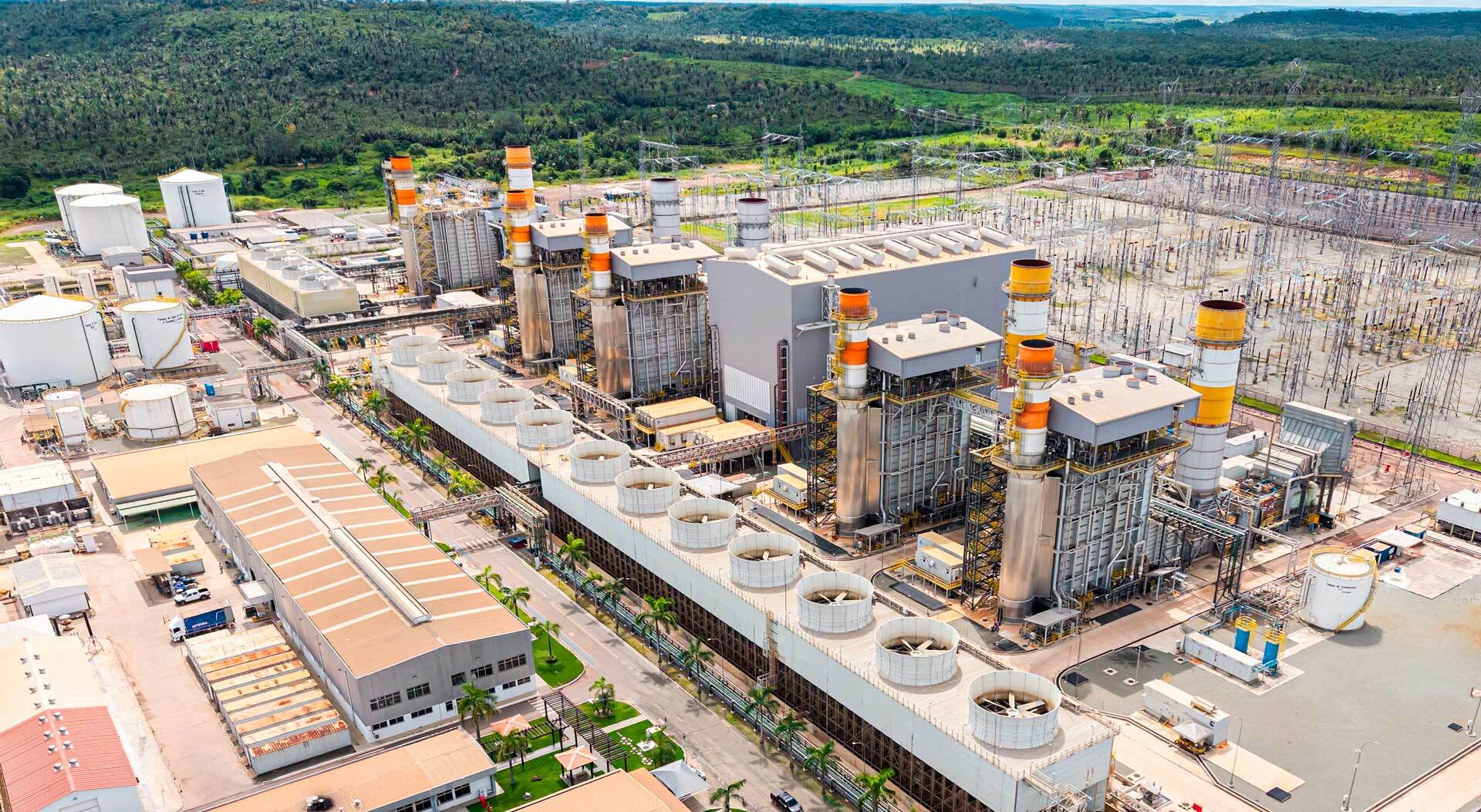Vista aérea de uma usina termelétrica, com turbinas e chaminés industriais de grande porte, tanques de armazenamento e uma subestação elétrica ao lado