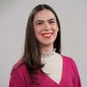 Retrato da jornalista Jamille Niero sorrindo, vestindo cardigan rosa e blusa off-white, contra um fundo cinza.